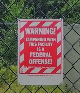 Red and white sign that reads: warning! Tampering this facility is a federal offense. US Code Title 42, Section 3*** (the section is blurry). Below that, it says to report suspicious activity to... but does not provide contact information